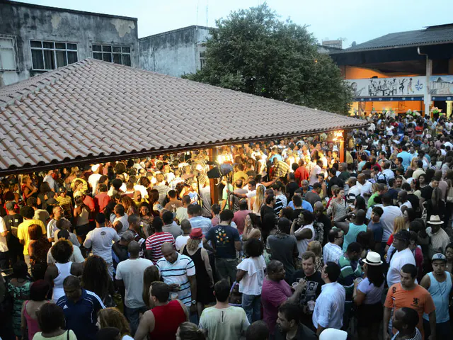 Crowd gathered under a large roof of tiles in an event at dusk, with buildings in the background