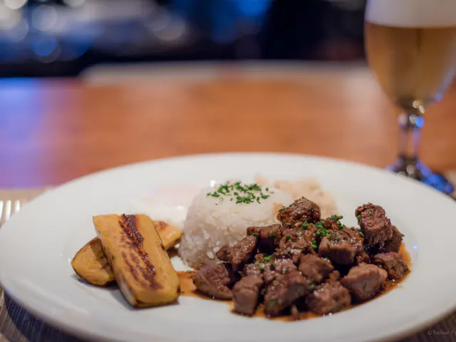 Platte mit Reis, Würfelfleisch und gebratener Banane, neben einem Glas Bier auf einem Holztisch