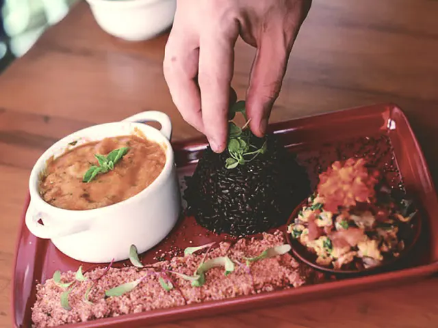 La mano termina con foglie verdi un piatto di moqueca con riso nero, foarofa e vinaigrette servito in vassoio rosso