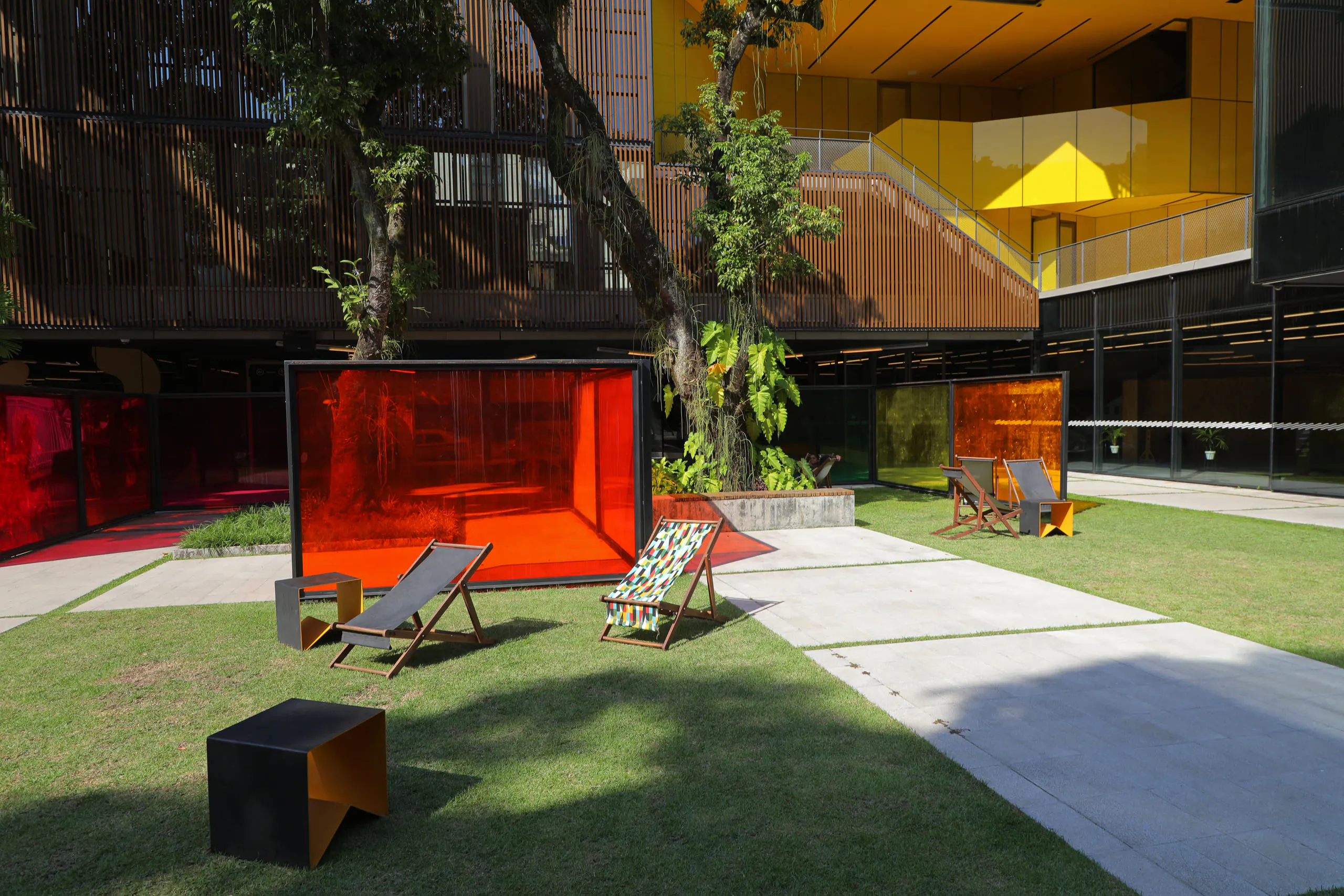Jardim gramado de edifício moderno, com espreguiçadeiras, um painel laranja e árvores diante de fachada com vidros amarelos