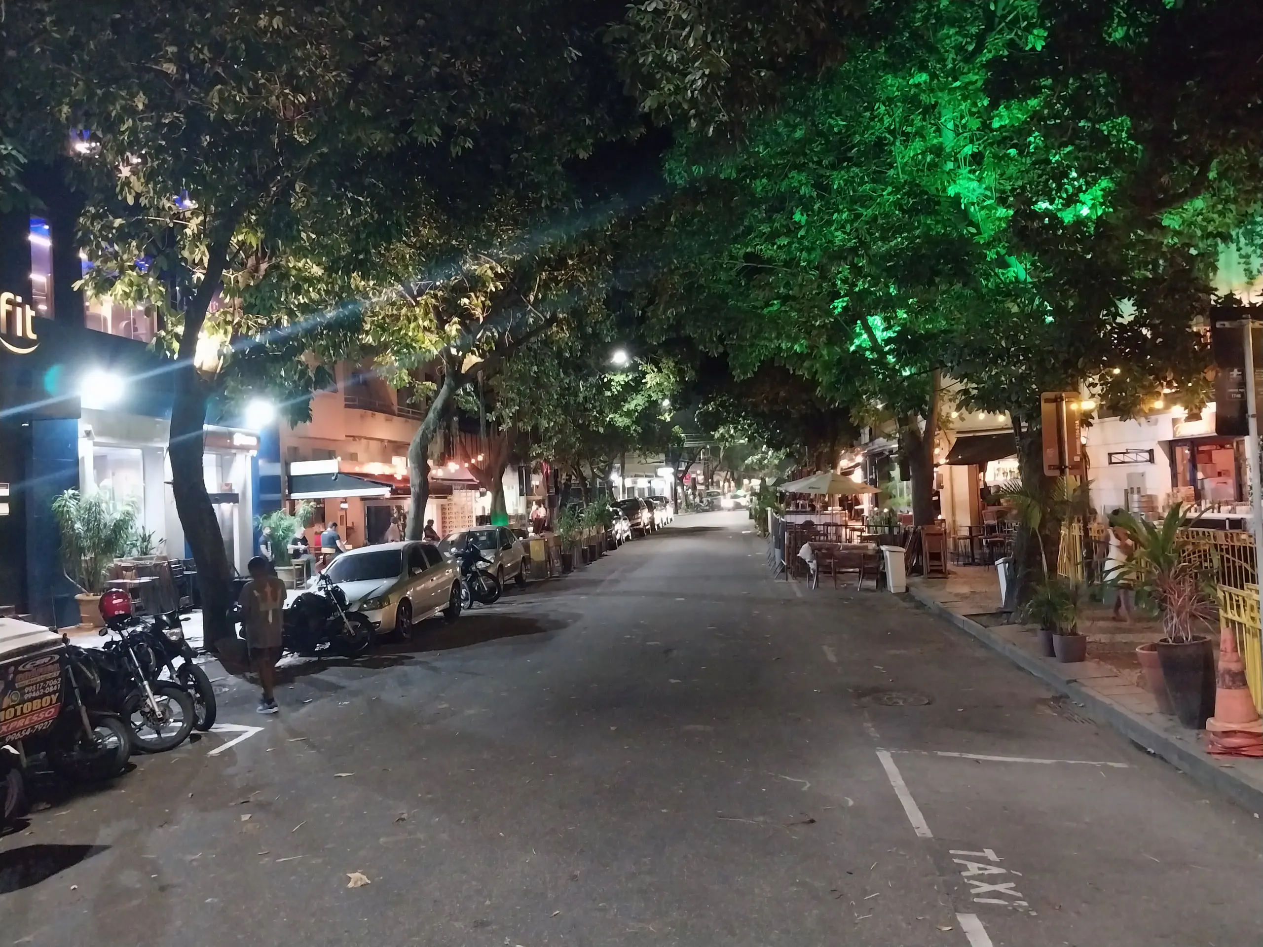 Rua de bairro à noite, com árvores iluminadas em verde, carros e motos estacionados e bares ao longo da calçada
